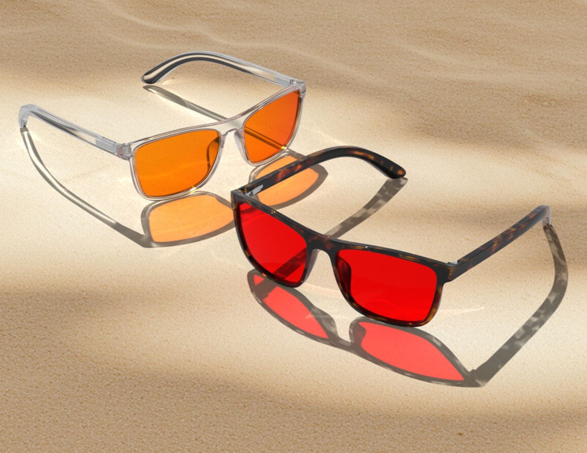 Two pairs of night ease sleep glasses with orange and red lenses rest on sunlit sand, casting colorful reflections