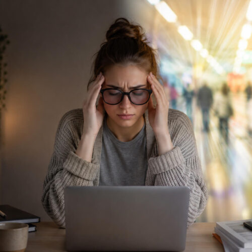Woman experiencing migraine and light sensitivity while using a laptop, illustrating sensory overload from bright indoor lighting and screens.