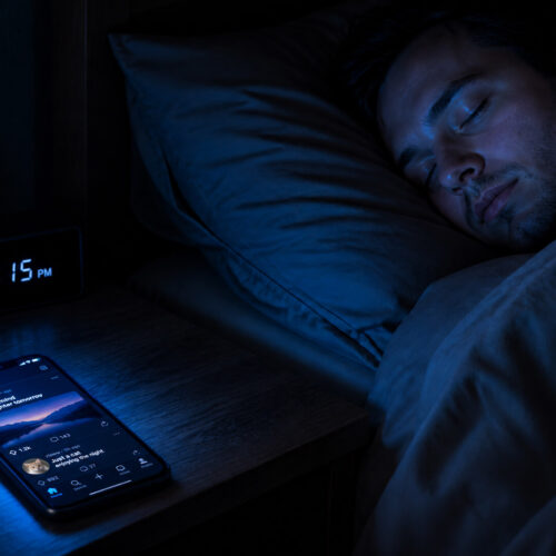 A smartphone glowing with cool blue light on a nightstand in a dark bedroom, its glow reaching a person lying in bed with eyes closed