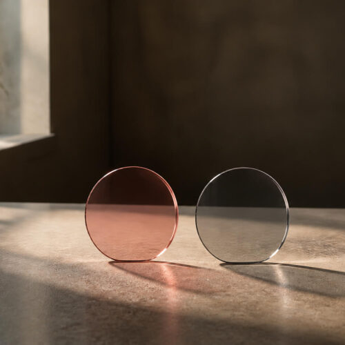 Two pieces of colored translucent lenses — rose and dark gray — placed side by side with soft window light passing through them, casting distinct colored shadows on a surface.