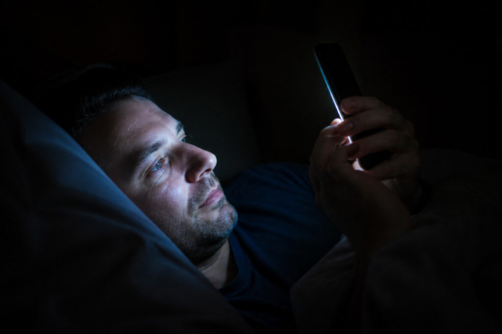Close view of a person lying in a dark bedroom, face and hands lit only by a smartphone screen